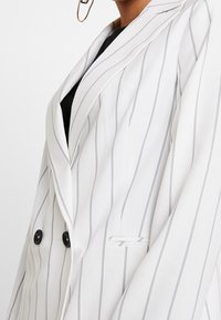 Valkoinen raidoitettu bleiseri, jossa on viimeistelty kaulus, kaksiriviset napit ja yksi tasku sivussa. Silkkinen kangas.