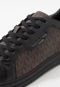 Svart sneakers med en texturerad brun tygöverkropp som har en logotyp, svarta snören och en slät svart läderdetalj nära hälen.