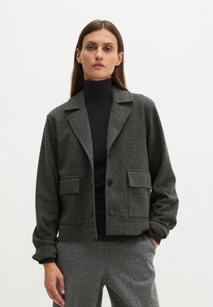 Vrouw draagt donkergrijze knoopjas met rever, bijpassende broek en zwarte coltrui tegen een effen lichte achtergrond.