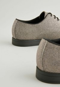 Glitzernde graue Schuhe mit glatter Textur, runder Zehenform, schwarzer Gummisohle und schwarzen Schnürsenkeln; Nähdetails entlang der Kanten.