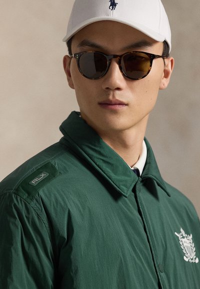 Mees kannab kilpkonnamustriga päikeseprille, valget pesapalli nokamütsi musta logoga ning rohelist jope, millel on valge tikitud vapp ja õlal RLX plaastriga.