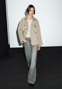 Trench coat beige doppiopetto, top rosa chiaro, jeans grigi a gamba larga, scarpe nere, in piedi contro uno sfondo bianco con un pavimento scuro.