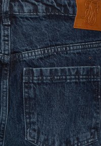Dark blue denim jeans featuring a back pocket and a tan leather patch with embossed initials. The texture is slightly faded and distressed.