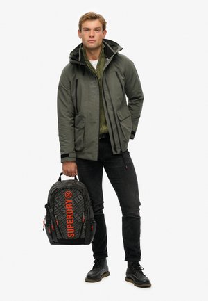Grøn vandtæt jakke med lynlåse, sorte jeans og en sort rygsæk med et gråt mønster og en lys orange logo, holdt i højre hånd.
