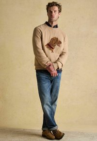 Kootud beež särk pruuni tikitud koera pea kujundusega, koos siniste teksade ja pruunide kingadega, seismas neutraalse tausta ees.