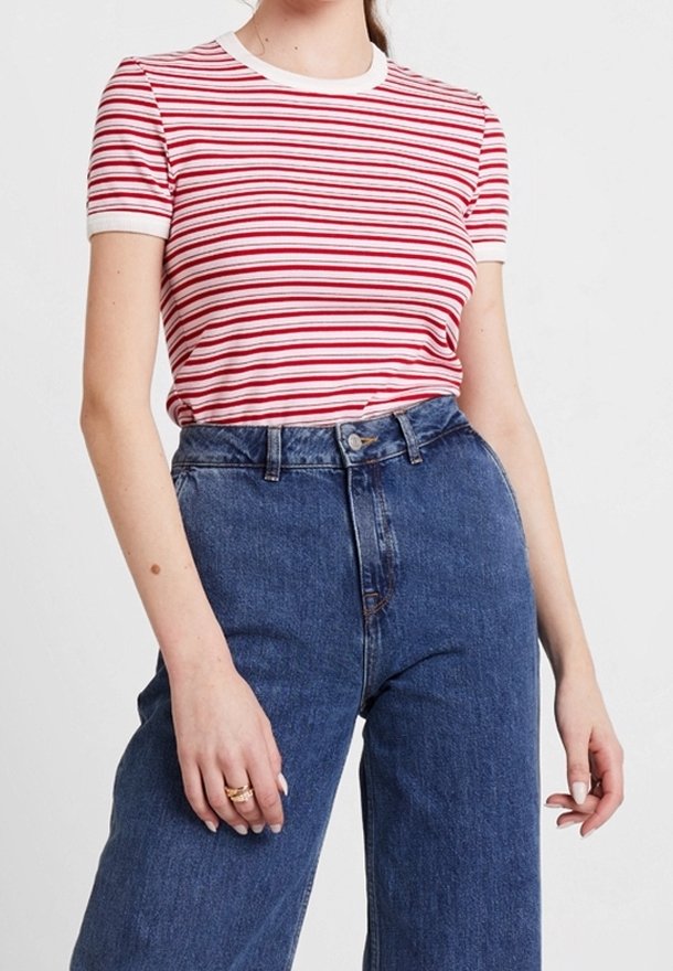 Femme portant un haut à manches courtes rayé rouge et blanc, rentré dans un jean en denim bleu taille haute, avec des bagues à la main gauche.