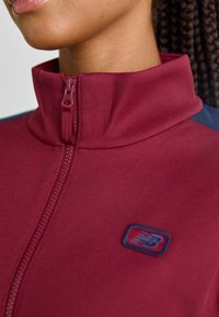 Bordeaux zip-jakke med høj krave og blå accenter. Har tekstureret stof og et lille logo nær lynlåsen.