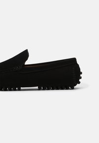 Mocassin en daim noir avec des clous ronds en caoutchouc sur la semelle et le talon, design slip-on à coupe basse et coutures apparentes.