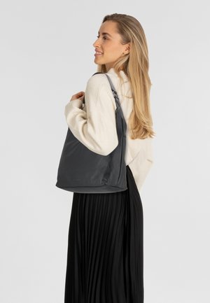 Junge Frau mit langen blonden Haaren, die einen cremefarbenen Pullover und einen schwarzen, plissierten Rock trägt und eine große schwarze Ledertasche über der Schulter hält.