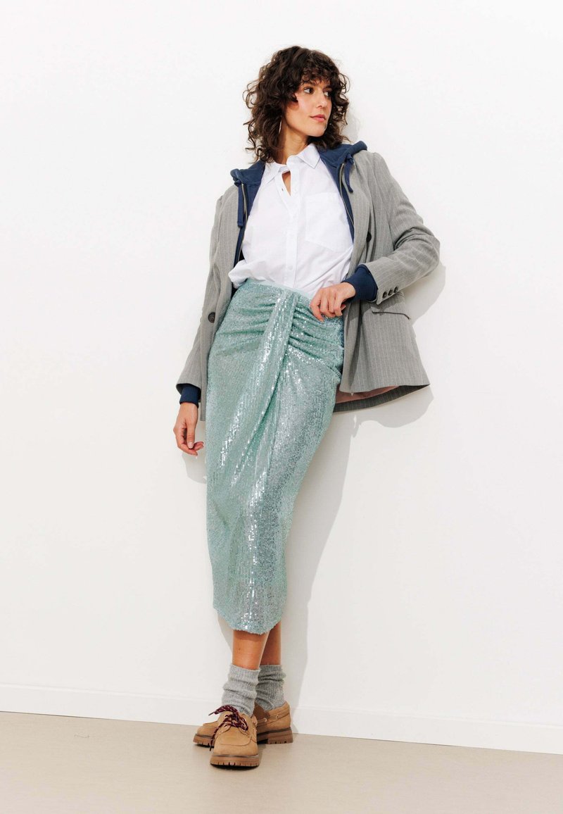 Jupe en sequins vert menthe métallique avec un design froncé, associée à un blazer gris sur une chemise blanche, des chaussures à lacets marron et des chaussettes grises.