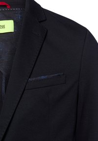 Schwarze Blazer mit Reverskragen, einer einzelnen Brusttasche mit marineblauem Akzent und einem strukturierten Innenfutter mit dezentem Muster.