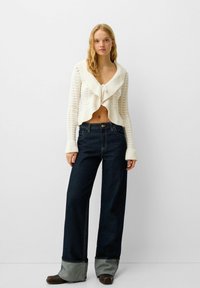 Cardigan court en tricot blanc avec des poignets côtelés, sur un jean large en denim foncé taille haute avec un ourlet gris replié. Fini lisse.
