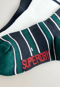 Stribede sokker i marineblå, grønne og hvide farver med rød "SUPERDRY" branding ved manchetten. Lavet af blødt, tekstureret stof.