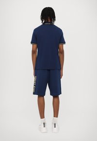 Mørkeblå polo med korte ermer, med kontraststriper i gult. Kombinert med matchende mørkeblå shorts som viser "VERSACE JEANS" i gull.