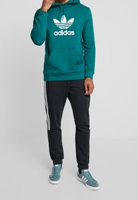 Person som bär en teal Adidas hoodie, svarta Adidas träningsbyxor med vita ränder och teal Adidas sneakers, står framför en grå bakgrund.