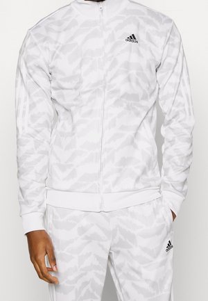 Weißer Adidas-Trainingsanzug mit hellgrauem Camouflage-Muster, Reißverschlussjacke und passenden Hosen, mit schwarzen Adidas-Logos auf Brust und Oberschenkel.