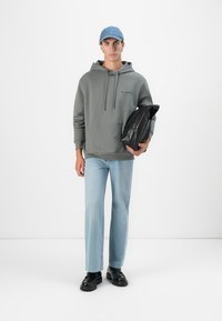 Mann iført grå hettegenser, lyseblå jeans og blå caps, holder en svart sammenfoldet jakke mot en hvit bakgrunn.
