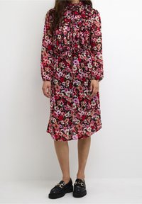 Kukkamekko, jossa on korkea pääntie ja pitkät hihat, valmistettu kevyestä kankaasta punaisen, vaaleanpunaisen ja mustan sävyissä, jossa on röyhelöyksityiskohtia ja nauhat.