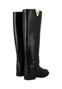 Bottes noires en cuir montantes avec une semelle plate, texture lisse et un accent doré en forme de V en haut. Détail de fermeture éclair sur le côté.