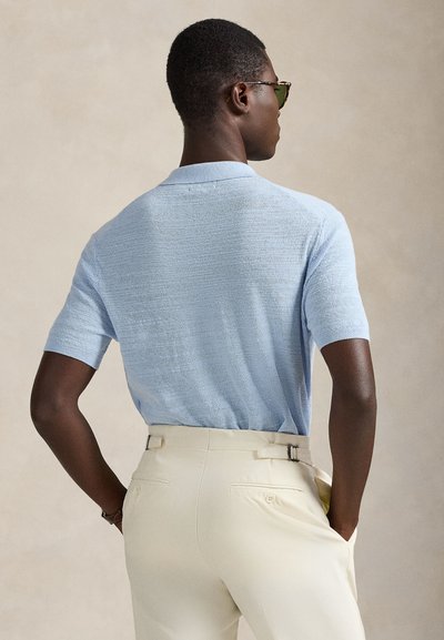 Πλεκτό πόλο μπλουζάκι σε ανοιχτό μπλε χρώμα με κοντά μανίκια, διαθέτοντας υφή στον ύφασμα. Συνδυασμένο με κρεμ παντελόνια που έχουν διπλές θηλιές για ζώνη.