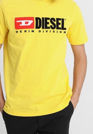 T-shirt en coton jaune avec un logo "DIESEL" noir et rouge et le texte "DENIM DIVISION" en dessous. Manches courtes et col rond.