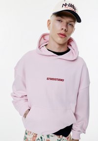 Ljusrosa hoodie med känguruficka och "STWDSTUDIO"-logotyp i vinröd. Avslappnad passform med dragsko på huvan, matchad med mönstrade byxor.