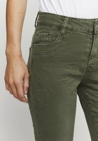 Pantalones de mezclilla de color verde oliva con cintura alta, cinco bolsillos y una sutil textura. Incluye trabillas para cinturón y cierre con cremallera.