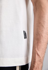 T-shirt bianco in cotone con maniche corte, dotato di un'etichetta nera sul lato con testo bianco. Tessuto liscio e vestibilità morbida.