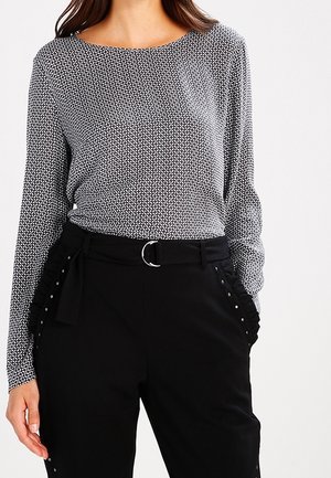 Kvinde iført en mønstret langærmet top stoppet ned i højtaljede sorte bukser med et stofbælte og flæse- og nitte-detaljer ved lommerne.