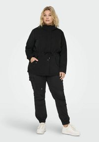 Juodas švarkas su reguliuojamu liemeniu, dviem priekinėmis kišenėmis ir aukštu apykakle, derinamas su juodomis kargo kelnėmis ir baltais sportbačiais. Paprastas dizainas.