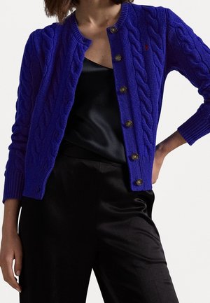 Kvinde iført en blå kabelstrikket cardigan med knapper over en sort satin top og sorte bukser mod en hvid baggrund.