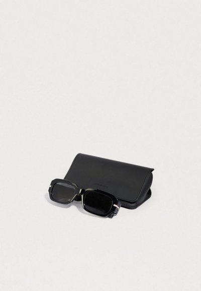 Fekete napszemüveg négyzet alakú kerettel, ezüst részletekkel, egy sima felületű fekete bőr tokkal mellett elhelyezve.
