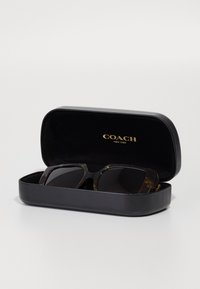 Musta kõva ümbris, millel on kuldne "COACH NEW YORK" logo, ja milles on üle mõõtmete prillid, millel on kilpkonnakujuline raam ja tumedad objektiivid.