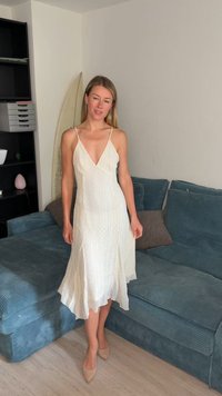 Femme en robe blanche texturée sans manches et talons beiges debout près d'un canapé bleu dans un salon avec des étagères et une planche de surf.