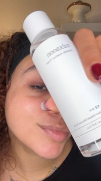 Heldere glazen fles met een witte dop, gelabeld "mixsoon", dicht bij een gezicht gehouden. De fles bevat een heldere vloeistof. Op de achtergrond is een plank met een keramisch voorwerp te zien.