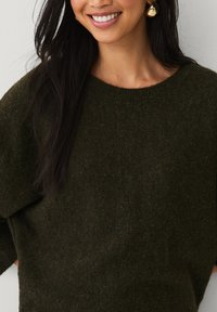 Pull vert foncé avec une texture douce, col large et coupe décontractée. Des boucles d'oreilles en or sont visibles à côté d'un visage souriant et de cheveux foncés.