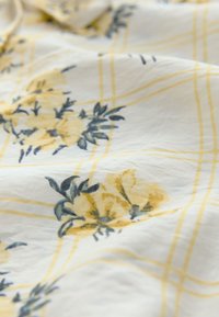 Tessuto floreale con fiori gialli e foglie verdi su sfondo bianco, sovrapposto da un sottile motivo a griglia gialla. Tessuto con trama stropicciata.