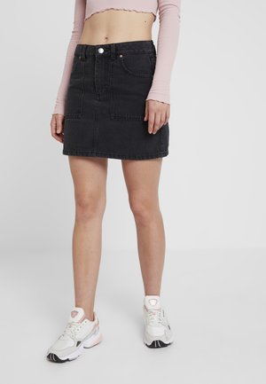 Femme portant une mini-jupe en jean noire avec des poches avant, un haut rose court à manches longues et des baskets blanches avec des accents roses.