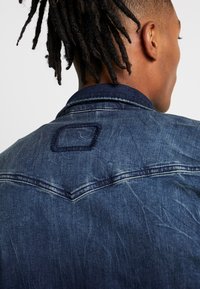 Giacca di jeans blu scuro con un taglio classico, cuciture visibili e una toppa quadrata sul retro. Leggera texture con un aspetto consumato.