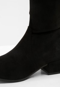 Gros plan sur une botte de cheville en daim noir avec un petit talon carré et des coutures visibles sur fond blanc.