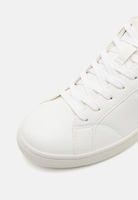 Sapatilha de pele branca com biqueira arredondada, superfície texturizada, atacadores brancos planos e costura tonal; sola de borracha com aderência texturizada.