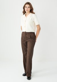 Korte mouw crèmekleurige gebreide cardigan met knopen en een borstzak, gecombineerd met hoog-waisted bruine patroonbroek en zwarte schoenen.