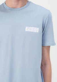 Helles blaues Baumwoll-T-Shirt mit kurzen Ärmeln, das auf der linken Brustseite ein weißes rechteckiges "GUESS"-Logo aufweist. Glatte Textur.