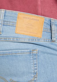 Jeans din denim albastru deschis cu o etichetă din piele maro, având text embosat. Cusături detaliate și o ușoară decolorare pe țesătură.