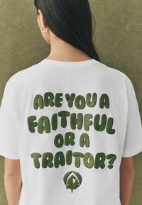 Hvid T-shirt med fed, grøn tekst der lyder "ER DU TROFAST ELLER EN FORRÆDER?" og en lille grafik nedenfor. Simpelt rundhalset design.