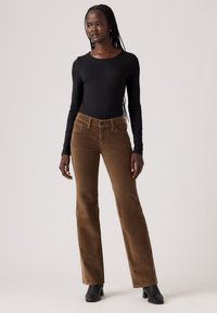 Bruine fluwelen broek met uitlopende pijpen en een gestructureerde afwerking, gecombineerd met een strak zwart langemouwshirt. Zwarte enkelboots maken de look compleet.