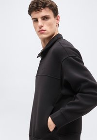 Νεαρός άνδρας με καστανά μαλλιά, φοράει μαύρο φούτερ με φερμουάρ μέχρι τη μέση, με το χέρι στην τσέπη, κοιτάζοντας προς την κάμερα.