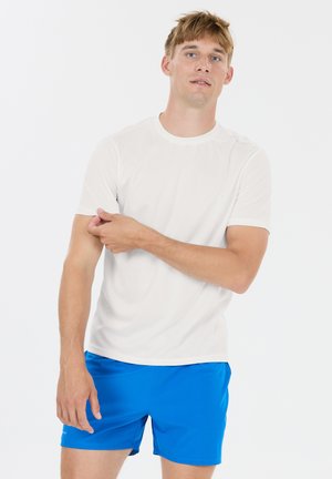 Ung mand, der retter ærmet på hvid T-shirt, iført lyseblå shorts, stående foran en ensfarvet hvid baggrund.