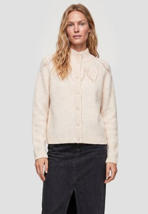 Kvinde med bølget blondt hår iført en cremet knapstrikket cardigan med bladmønstre og et mørkt knælængde skørt med en frontslids.