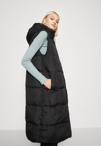 Zwarte puffer vest met een capuchon, met een lange, gebatteerde uitstraling, zijzakken en een gladde textuur. Draag het over een lichtblauwe geribbelde top.
